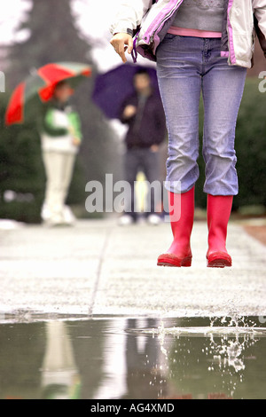 Jeune femme sauter dans une flaque d'eau avec des amis en arrière-plan l'exercice parapluies dans la pluie 12 29 ans 20 ans 20 s 20 30 Banque D'Images