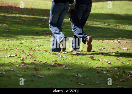 Moitié inférieure d'un couple portant des jeans de marcher à travers l'herbe avec les feuilles d'automne dans un parc public Banque D'Images