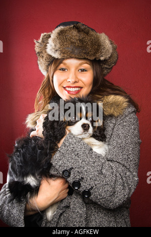 Young adult Caucasian woman wearing fur hat holding King Charles Spaniel dans arms and smiling at viewer Banque D'Images