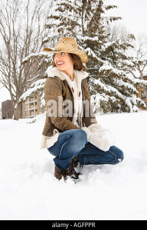 Portrait jeune femme adulte à plus d'épaule, tout en s'agenouillant dans la neige avec boule de neige et wearing straw cowboy hat Banque D'Images