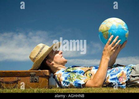 Jeune homme sur fond de ciel bleu portant des vêtements de vacances chemise hawaïenne sandales short straw hat contemplant globe world Banque D'Images