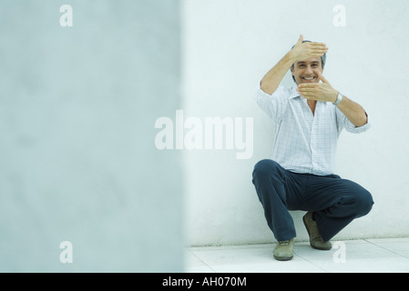 Man crouching, encadrant le visage avec les mains, smiling Banque D'Images