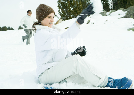 Teenage girl sitting on ground in snow, lancer snowball, side view Banque D'Images