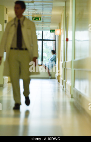 Homme assis, looking out window, portant chemise d'hôpital, médecin en premier plan de marche Banque D'Images