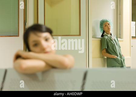 Doctor standing with hands in pockets, looking at camera, woman resting head on arms en premier plan Banque D'Images