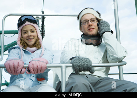 Young man using cell phone, assis sur une chaise ascenseur avec soeur, tous deux habillés en vêtements d'hiver Banque D'Images