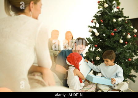 L'homme et deux enfants opening Christmas present in front of Christmas Tree, femme de regarder en premier plan Banque D'Images