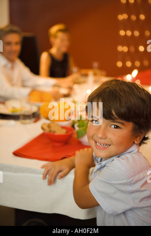 Garçon assis à la table du dîner, smiling at camera, adultes en arrière-plan Banque D'Images