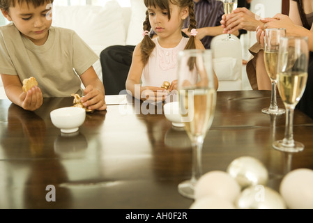 Garçon et fille assis à table, les deux holding Christmas Ornaments, adultes de boire du champagne en arrière-plan Banque D'Images