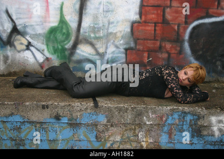 Une femme aux cheveux blonds porter des pantalons noirs et une blouse noir est posé sur une corniche en brique en face d'un mur de briques graffited Banque D'Images