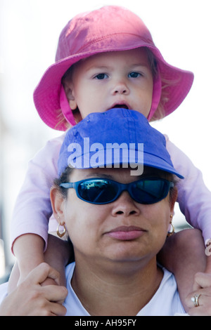 Fillette de deux ans dans un chapeau rose assis sur les épaules de sa mère qui porte un chapeau et des lunettes de soleil violet Banque D'Images