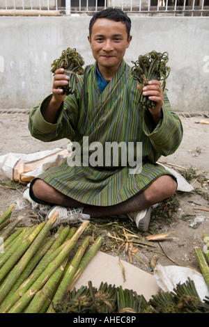 Heureux jeunes Bhoutanais sitting cross legged et portant un gho propose des pousses de fougère Osmunda comestibles et de la canne à sucre à vendre . Banque D'Images