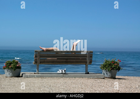 Une femme lit un livre dans une position délicate sur un banc avec vue sur l'océan. Banque D'Images