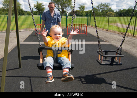 Close up of a horizontal 16 mois baby boy clapping avec enthousiasme comme il a poussé sur une balançoire par son père à un jeu pour enfants Banque D'Images