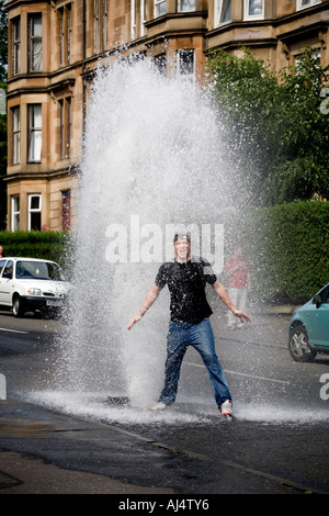 Un jeune homme se refroidit dans la chaleur de l'été comme un poteau de l'eau principale burst et jaillit de l'eau dans une rue de Glasgow Dennistoun UK Banque D'Images