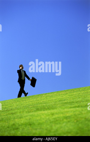 Man in suit with briefcase walking up grassy hill Banque D'Images