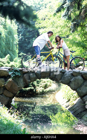 Jeune couple riding bikes sur un pont en pierre dans un parc. Banque D'Images