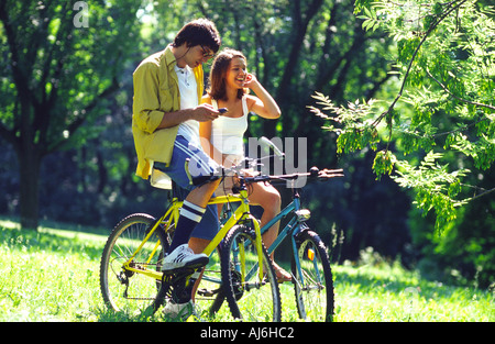 Jeune couple riding a vélos dans le parc et en utilisant les téléphones mobiles. Banque D'Images