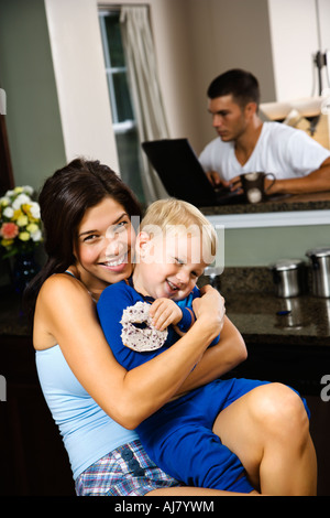 Caucasian woman hugging petit garçon dans la cuisine avec le père sur l'ordinateur portable en arrière-plan Banque D'Images