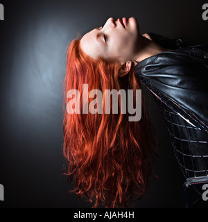 Jolie rousse jeune femme portant veste en cuir se penchant en arrière dans un fauteuil avec les cheveux longs pendant vers le bas Banque D'Images