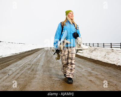 Jeune femme dans des vêtements d'hiver marchant dans un chemin de terre boueux holding snowboard et boots smiling Banque D'Images