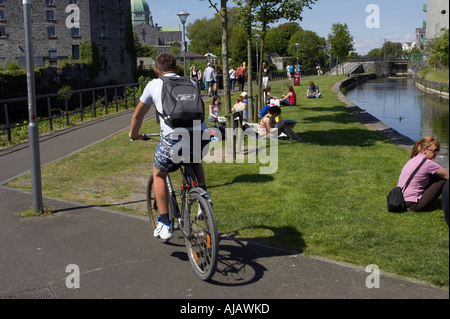 Vélo homme passé, les gens se détendre au soleil sur l'herbe à côté du chemin de halage d'un canal Banque D'Images