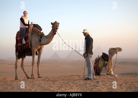 Touriste assis sur un chameau avec guide dans le désert pierreux dans la lumière du soleil tôt le matin brumeux misty avec pyramides dans la distance Banque D'Images