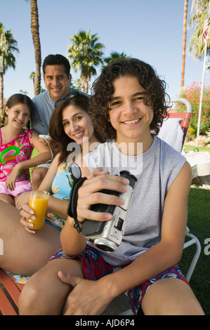 Boy (13-15) holding caméscope, assis à l'extérieur avec sa sœur (7-9) et les parents, portrait. Banque D'Images