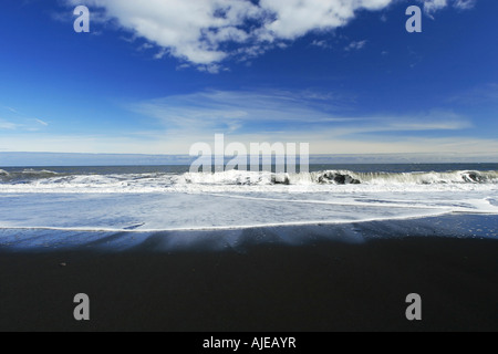 Vagues se brisant sur une plage de sable noir fond bleu ciel et nuages en arrière-plan Banque D'Images