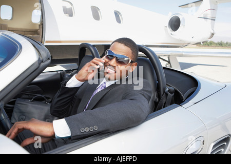 Man sitting in Convertible près de jet privé, talking on mobile Banque D'Images