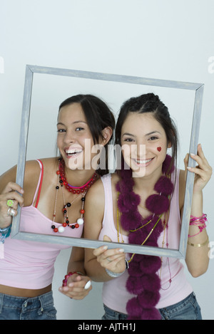 Deux jeunes femmes friends holding up photo frame, smiling at camera, portrait Banque D'Images