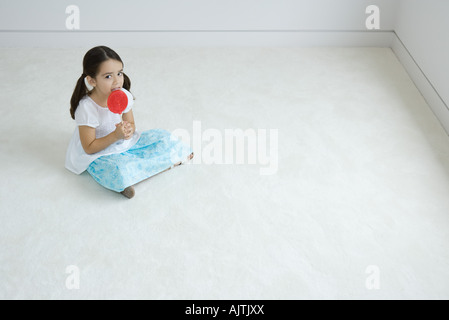 Petite fille assise sur le plancher, le léchage grand lollipop Banque D'Images