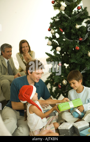 Le père et les deux enfants assis par arbre de Noël, l'ouverture présente ensemble, les adultes en arrière-plan Banque D'Images