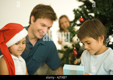 Le père et les deux enfants assis par arbre de Noël, daughter wearing Santa hat, fils présents ouverture Banque D'Images
