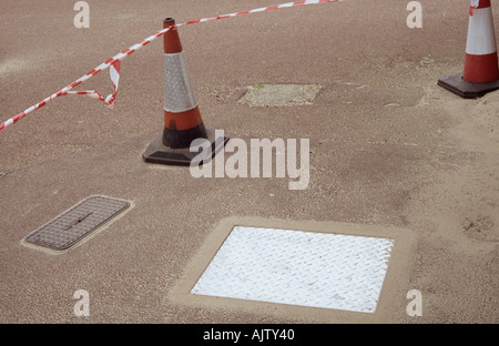 Un couvercle de trou d'aluminium qui vient sur un trottoir avec des correctifs et des bandes cônes de travaux Banque D'Images