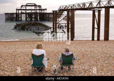 Un homme âgé et une femme et son chien assis sur la plage par le reste de la jetée Ouest, Brighton, Angleterre. Banque D'Images