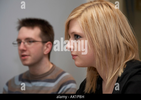 Jeune femme aux cheveux blond assis à côté de dark haired young man wearing glasses entendre quelqu'un Banque D'Images