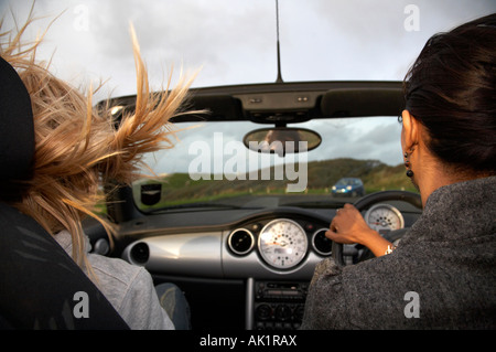 Young Asian woman au volant d'une mini cabriolet le long de la mer dans la soirée à la vitesse avec fille irlandaise blond Banque D'Images