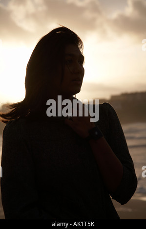 Silhouette de dark haired asian female student standing sur une plage au coucher du soleil face à la mer à l'aide jusqu'à son cou Banque D'Images