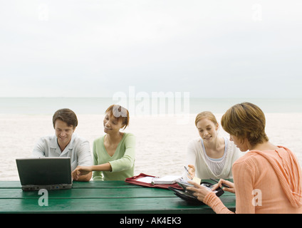 Quatre adultes assis à table de pique-nique sur la plage, looking at laptop et ordres du jour Banque D'Images