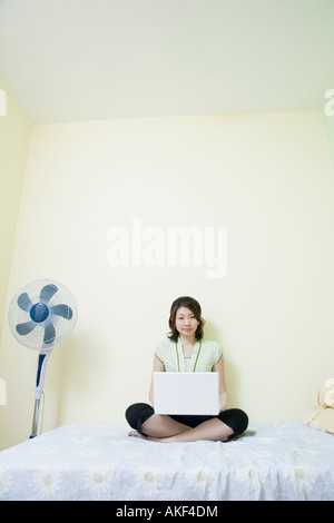 Jeune femme assise en tailleur sur le lit et à l'aide d'un ordinateur portable Banque D'Images