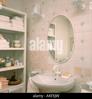 Miroir ovale sur rose et blanc mur au-dessus de bassin dans une petite salle de bains avec serviettes et articles de toilette sur l'étagère en rotin blanc Banque D'Images