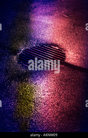 Qui se reflète sur les néons de la chaussée mouillée et d'égout à grille de vidange Banque D'Images