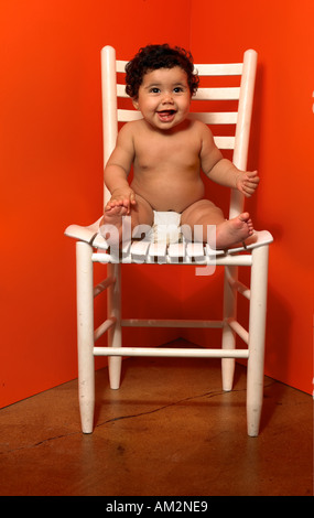 Un mignon bébé rit alors qu'il était assis sur une chaise par elle-même contre l'angle d'un mur orange Banque D'Images