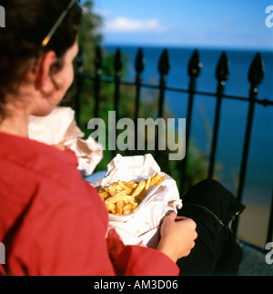 Arrière arrière arrière vue jeune femme mangeant du poisson et des frites Situé au-dessus de South Beach sur la côte galloise Tenby Pembrokeshire Pays de Galles Royaume-Uni Grande-Bretagne KATHY DEWITT Banque D'Images