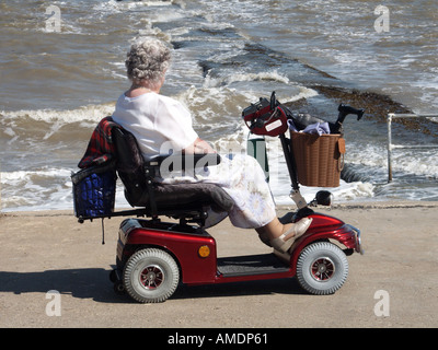 Front de mer promenade de mur de mer femme mature conduisant sur scooter d'invalidité motorisée regardant à marée haute mer rugueuse Walton sur le Naze Essex Angleterre Royaume-Uni Banque D'Images