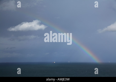 Un arc-en-ciel vif voûtant au-dessus d'un océan bleu foncé avec un ciel nuageux et des navires lointains Banque D'Images