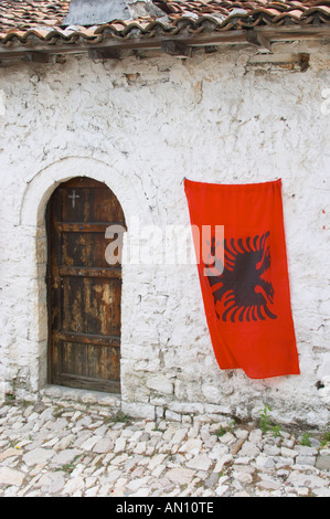 Le drapeau albanais, rouge avec double tête noire blanche, contre un mur de pierres blanches. Vieille porte en bois. Berat la citadelle vieille ville fortifiée. L'Albanie, des Balkans, de l'Europe. Banque D'Images