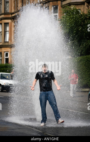 Un jeune homme se refroidit dans la chaleur de l'été comme un poteau de l'eau principale burst et jaillit de l'eau dans une rue de Glasgow Dennistoun UK Banque D'Images