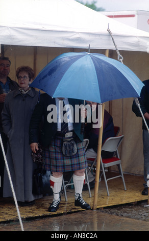 L'homme en kilt abrite de pluie pendant la coupe du Shinty match final contre Oban Newtonmore Banque D'Images